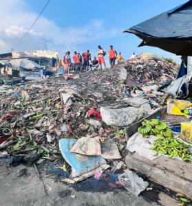 Group of people in orange safety vests standing on a large pile of trash at a dumpsite under a blue sky, with banana bunches visible on the side.