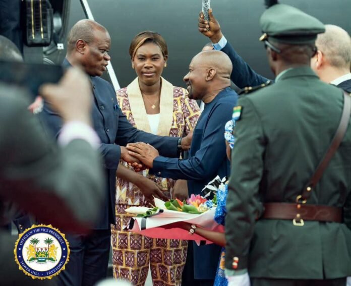 Two men shake hands on a tarmac while a woman in a patterned suit and others look on; a bouquet is being presented.