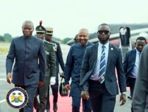 Dignitaries in dark suits walk on a red carpet at an airport, with a ceremonial guard and airplane in the background; official seal visible in the foreground.