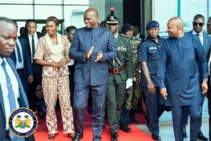 Group of dignitaries and security personnel walking onto a red carpet outside a building, central figure clapping.