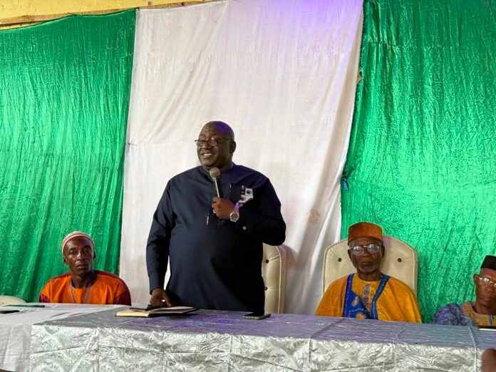 Man in dark traditional suit stands at a panel desk, speaking into a microphone as others in colorful attire sit nearby with a green and white backdrop behind them.