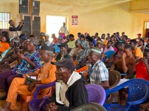 A large group of people seated in a yellow-walled hall listening to a community meeting; a photographer stands left taking pictures.