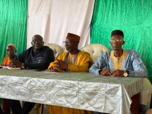 | The Calabash Newspaper Panel of four men seated at a table during a formal event, wearing traditional and modern attire, under a green and white tent backdrop.