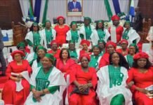 THE PRESIDENT WHO IS CHANGING SIERRA LEONE’S POLITICAL STORY Group of people in ceremonial attire posing for a photo in a formal hall; many wear red dresses and green-and-white traditional outfits, with a portrait on stage behind them.