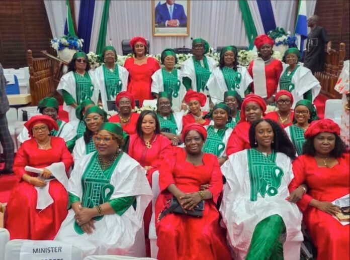 Group of people in ceremonial attire posing for a photo in a formal hall; many wear red dresses and green-and-white traditional outfits, with a portrait on stage behind them.
