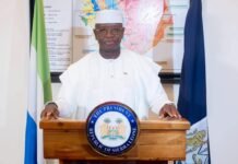 President Bio Urges Parliament to Vote “Yes” on Constitutional Amendment Bill Man in white traditional attire stands behind a wooden podium with the seal reading 'The President, Republic of Sierra Leone', flanked by flags.