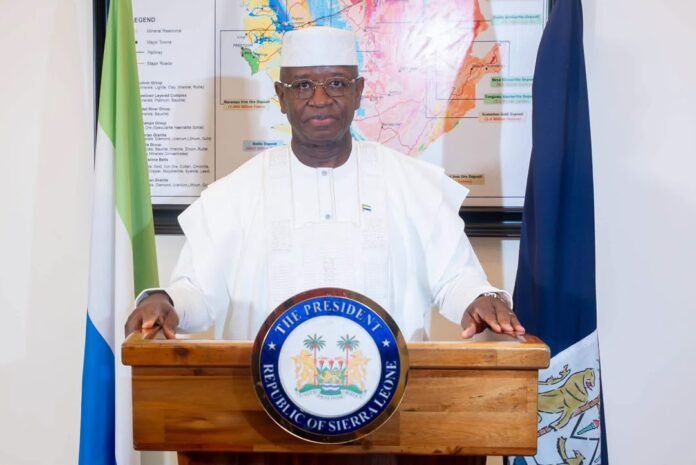 Man in white traditional attire stands behind a wooden podium with the seal reading 'The President, Republic of Sierra Leone', flanked by flags.