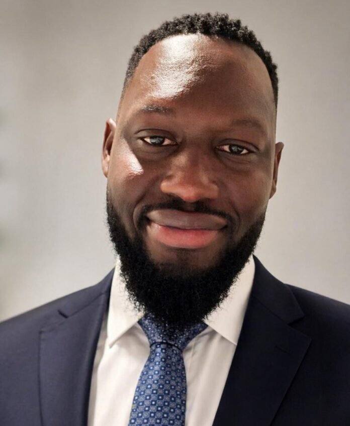 Professional headshot of a smiling man in a navy suit, white shirt, and blue patterned tie against a neutral background.
