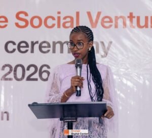 | The Calabash Newspaper Woman presenter in lavender lace dress speaking into a microphone at a lectern, with an Orange Digital Center banner in the background.