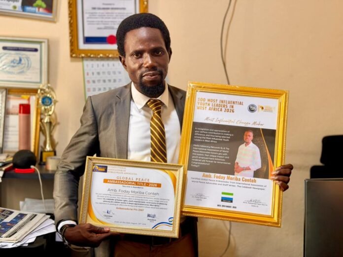 Man in a gray suit holds two gold-framed awards in an office setting, smiling at the camera.