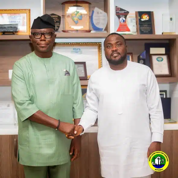 Two men in traditional outfits shake hands in an office with awards and certificates on shelves behind them.