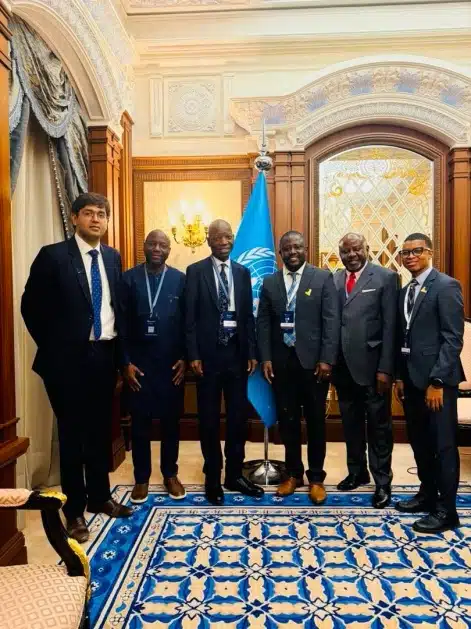 Six men in suits stand together in a gilded, ornate room with a United Nations flag behind them.