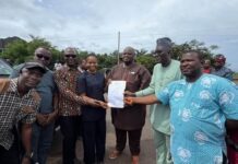 Lands Ministry Hands Over Land for Construction of Borbor Community Market Group of people outdoors presenting a document to a central man in brown, while others watch and smile near a parked car.