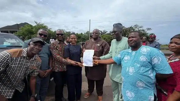 Group of people outdoors presenting a document to a central man in brown, while others watch and smile near a parked car.