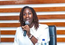 MCA-SL Sets September 2027 for Full Implementation of $480M Energy Compact Woman wearing a white blouse speaks into a handheld microphone at a panel discussion, sitting at a table with a water bottle nearby.