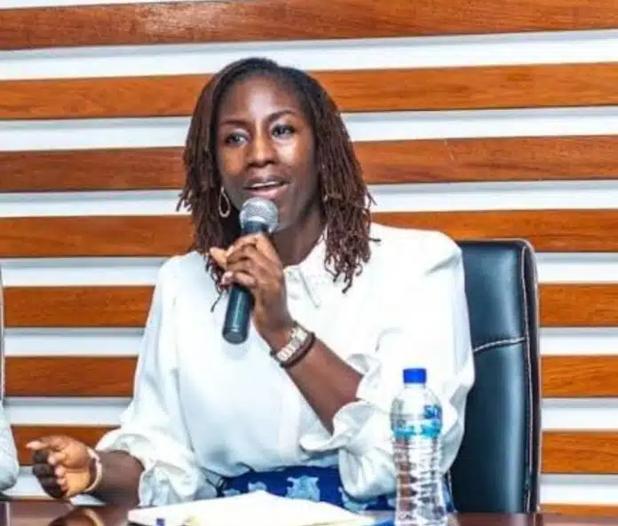 Woman wearing a white blouse speaks into a handheld microphone at a panel discussion, sitting at a table with a water bottle nearby.