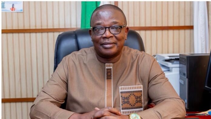 Portrait of a man wearing a brown traditional shirt and glasses, seated at an office desk and looking at the camera.