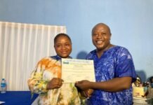 Make Impact, We Yone Child Foundation Equip 44 Vulnerable Youths with Vocational Skills Two people smile as one hands a certificate to the other during a small award ceremony in a blue-walled room with a table in the background.