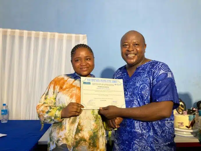 Two people smile as one hands a certificate to the other during a small award ceremony in a blue-walled room with a table in the background.