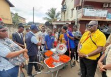Caritas Freetown Intensifies Flood Prevention with Three-Day Community & Main Street Cleaning Exercise Community volunteers gather on a street while a man in a blue vest explains the distribution of orange life jackets from a wheelbarrow toward a crowd of youths and adults.