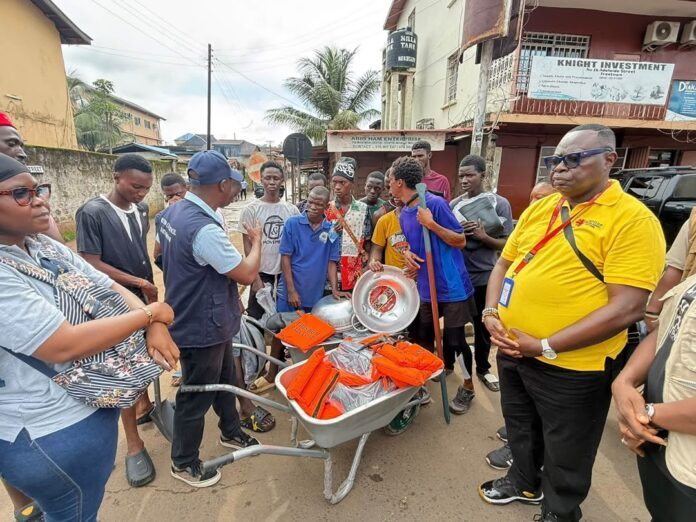 Community volunteers gather on a street while a man in a blue vest explains the distribution of orange life jackets from a wheelbarrow toward a crowd of youths and adults.