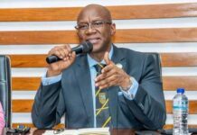 Mines Minister Unveils Mining Fiscal Framework Projecting US$3.63 Billion from Model Bauxite Mine Man in a dark suit and striped yellow tie speaks into a handheld microphone at a table during a meeting, with a notebook and water bottle nearby.