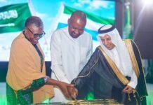 Vice President Jalloh Joins Saudi Community to Celebrate 96th National Day in Freetown Three dignitaries join hands to cut a ceremonial cake at a formal event, with green flags in the background.