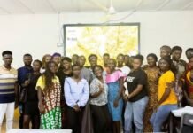 BBC Media Action, EU Empower University Students Through ‘Dis Na Wi Voice’ Project Group of people posing for a photo in a classroom or training room, with a large map projected on a screen behind them and a bright, well-lit setting.