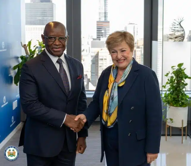 Two business professionals shake hands in a bright office with large windows and potted plants at the city skyline backdrop.