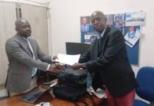 SLYDCL Policy Brief Highlights Gaps in Sierra Leone’s 2008 Drug Legislation Two men in business attire shake hands in an office as one passes a white document over the desk cluttered with a laptop and bags behind them.