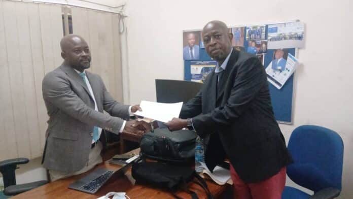 Two men in business attire shake hands in an office as one passes a white document over the desk cluttered with a laptop and bags behind them.
