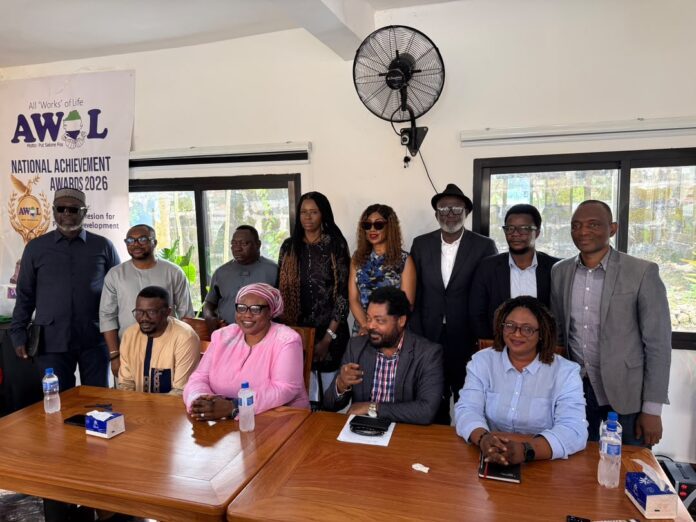 Group of fourteen people posing for a photo around a wooden conference table, with a banner reading National Achievement Awards 2026 in the background.