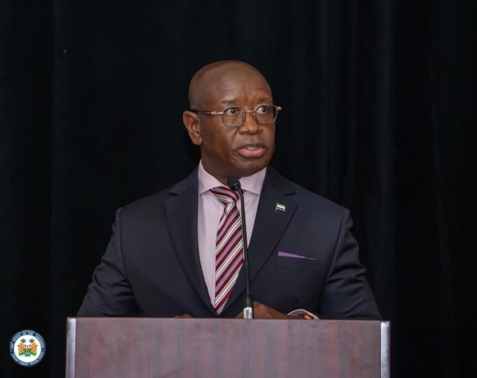 Professional man in a dark suit and striped tie speaks at a podium with a microphone, wearing glasses and a lapel flag pin, against a black backdrop.