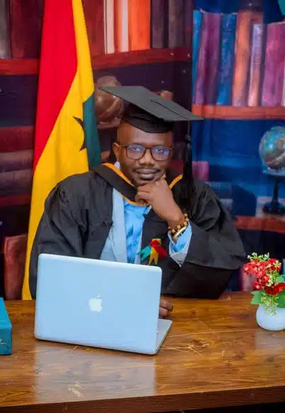 Graduate in cap and gown sits at a wooden desk with a laptop, flag and books in the background.