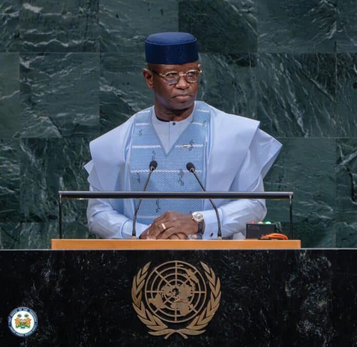 Man in light blue traditional attire and cap speaks at a UN podium with microphones and the UN emblem behind him.