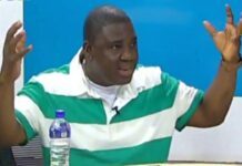 Moses Mambu’s Power-Drunk Rhetoric Risks Tearing the SLPP Apar Man in a green and white striped shirt raises both hands while speaking, with a water bottle on the table nearby.