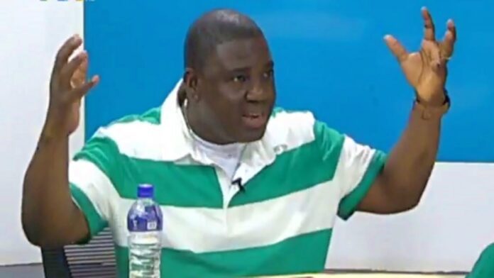 Man in a green and white striped shirt raises both hands while speaking, with a water bottle on the table nearby.