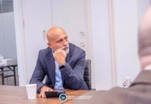 At UNGA… Africell Joins Government, Global Partners to Push Connectivity, 5G & Digital Infrastructure Investment A bald man in a blue suit sits at a conference table, resting his chin on his hand, with a tablet in front and a water bottle nearby.