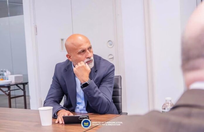 A bald man in a blue suit sits at a conference table, resting his chin on his hand, with a tablet in front and a water bottle nearby.