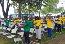 NP Sierra Leone Promotes Staff Wellness Through Monthly Health Walk Group of people in green and yellow shirts standing and stretching beside rows of white folding chairs at an outdoor event, with trees and a blue bus in the background.