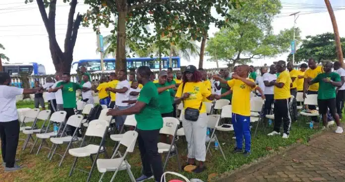 Group of people in green and yellow shirts standing and stretching beside rows of white folding chairs at an outdoor event, with trees and a blue bus in the background.