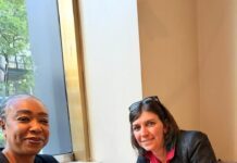 Peace Commission Executive Secretary Advances Sierra Leone’s Conflict Prevention Agenda During High-Level New York Engagements Two women sit at a wooden table by a window, smiling at the camera, with a stack of cards and a laptop on the table.