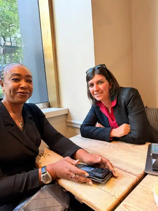 Two women sit at a wooden table by a window, smiling at the camera, with a stack of cards and a laptop on the table.