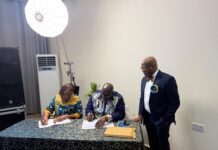 Institute of Directors Sierra Leone Relaunched, Signs MoU with Chamber of Commerce & Certifies 15 Directors Two people sign documents at a table while a suited man stands nearby in a studio setting with a large light in the corner.