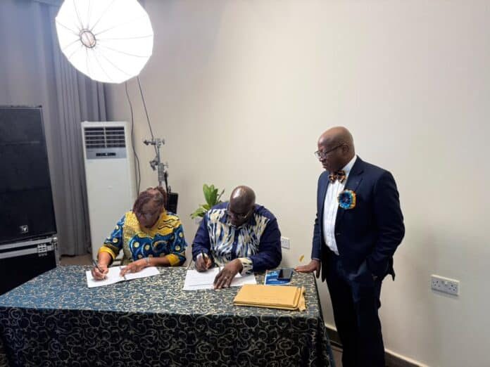 Two people sign documents at a table while a suited man stands nearby in a studio setting with a large light in the corner.