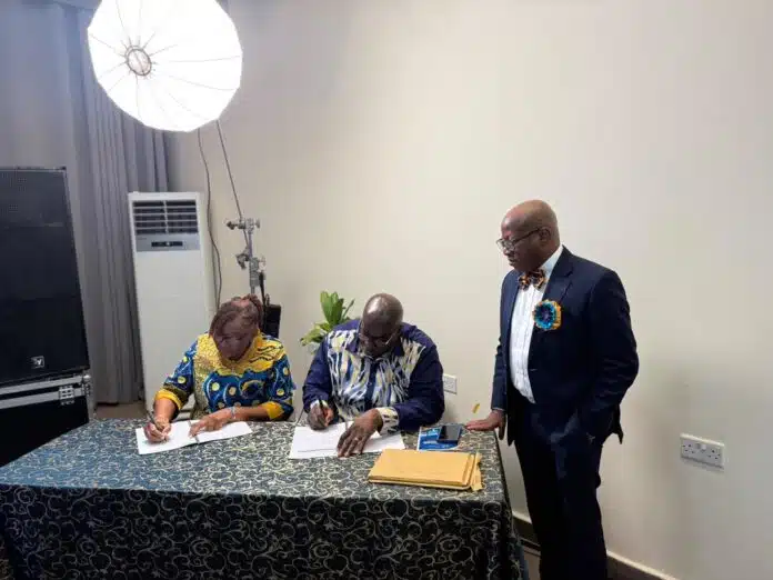 Two people sign documents at a table while a suited man stands nearby in a studio setting with a large light in the corner.