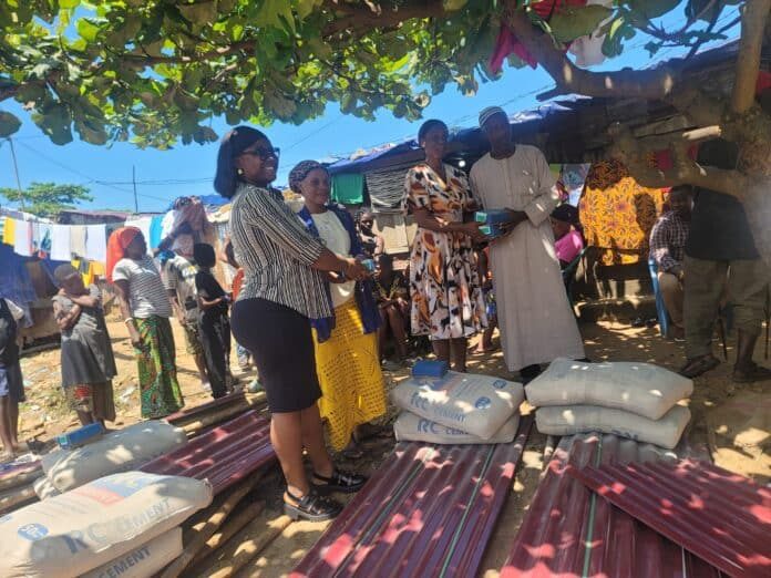 Group of people at an outdoor aid event under a leafy tree, exchanging a box while cement bags and metal roofing sheets lie nearby