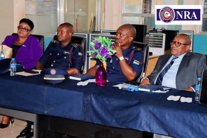 Panel of four people seated at a conference table; two in uniform, one woman in purple, one man in a suit, purple vase of flowers center stage; NRA logo in background.