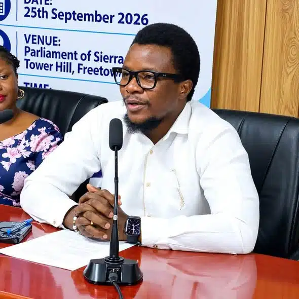 Man in a white shirt and glasses speaking into a microphone at a press conference, seated at a red table with notes; banner behind lists date and venue.