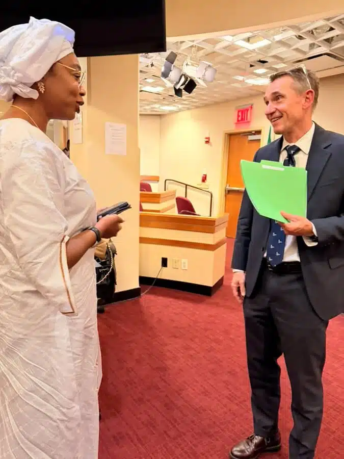 Two professionals chat in a hallway: a woman in white traditional attire holding a phone, facing a man in a suit with a bright green folder.